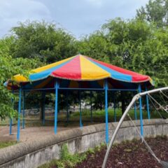 Open ronde tent met blauwe palen en geel-rood-blauw zeilen dak in boomrijke omgeving